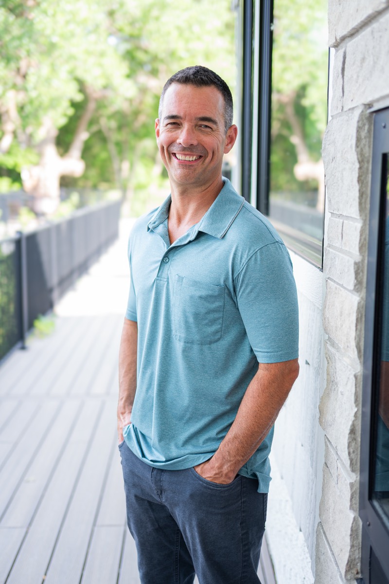 Amos Kittelson smiling in a teal polo, leaning against a stone wall